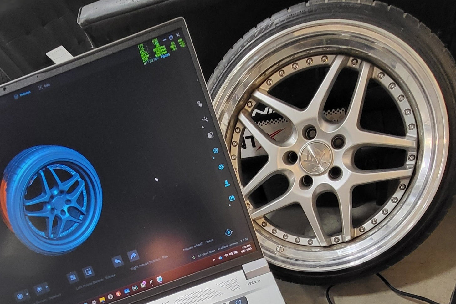 Laptop displaying a 3D model of a wheel next to a real wheel on a table. the wheel is being 3D scanned.